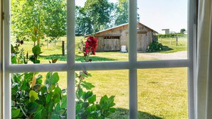 A view through a window with a wooden barn, green trees, and a grassy yard. - Holiday Home “La Petite Chaumière” with Shared Garden (Sainte-Foy)