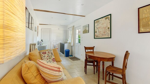 A living room with a sofa, a dining table with chairs, a kitchen in the background, and framed artwork on the walls. - Holiday Home “La Petite Chaumière” with Shared Garden (Sainte-Foy)