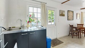 A kitchen with a sink, a window with a curtain, a dining area with chairs, and a door. - Holiday Home “La Petite Chaumière” with Shared Garden (Sainte-Foy)