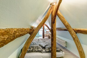 A loft bedroom with exposed wooden beams, a bed with patterned pillows, and a chair in the background.