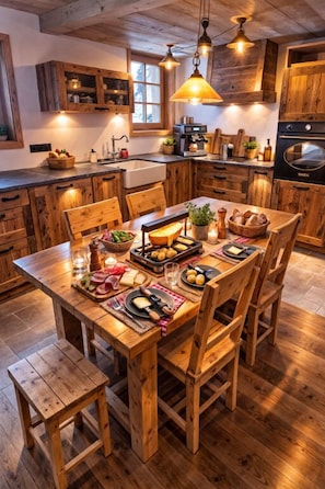 Una mesa de comedor de madera dispuesta con comida y bebidas, rodeada de sillas de madera, en una cocina con armarios de madera y una lámpara colgante.