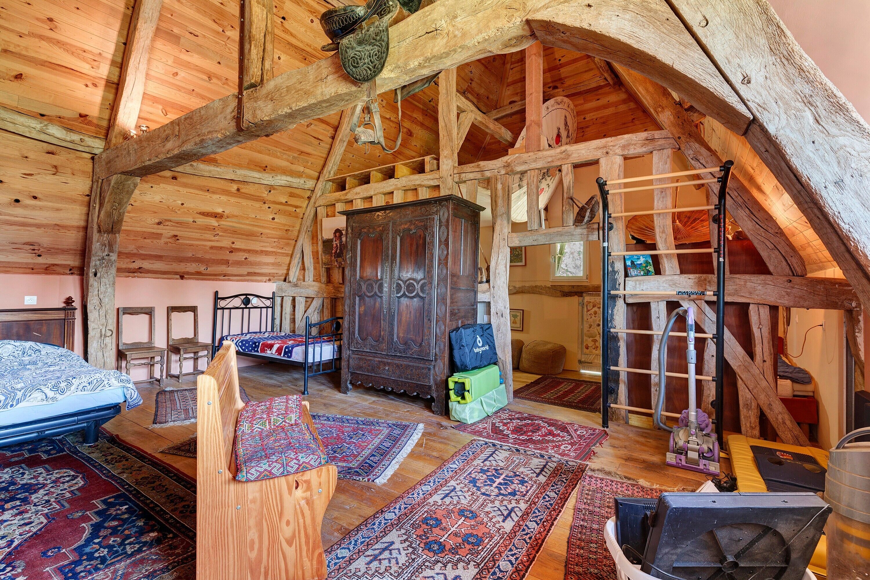 Une chambre mansardée avec des poutres apparentes, un lit, une armoire en bois, une échelle et un aspirateur.