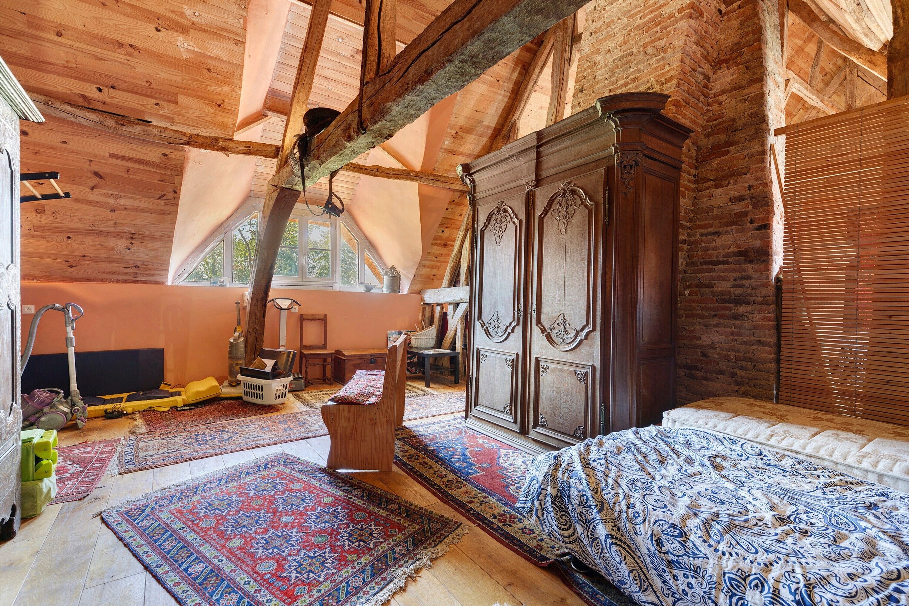 Una habitació amb sostre inclinat, bigues de fusta, un armari gran, un llit amb roba de llit estampada i un terra amb alfabres.