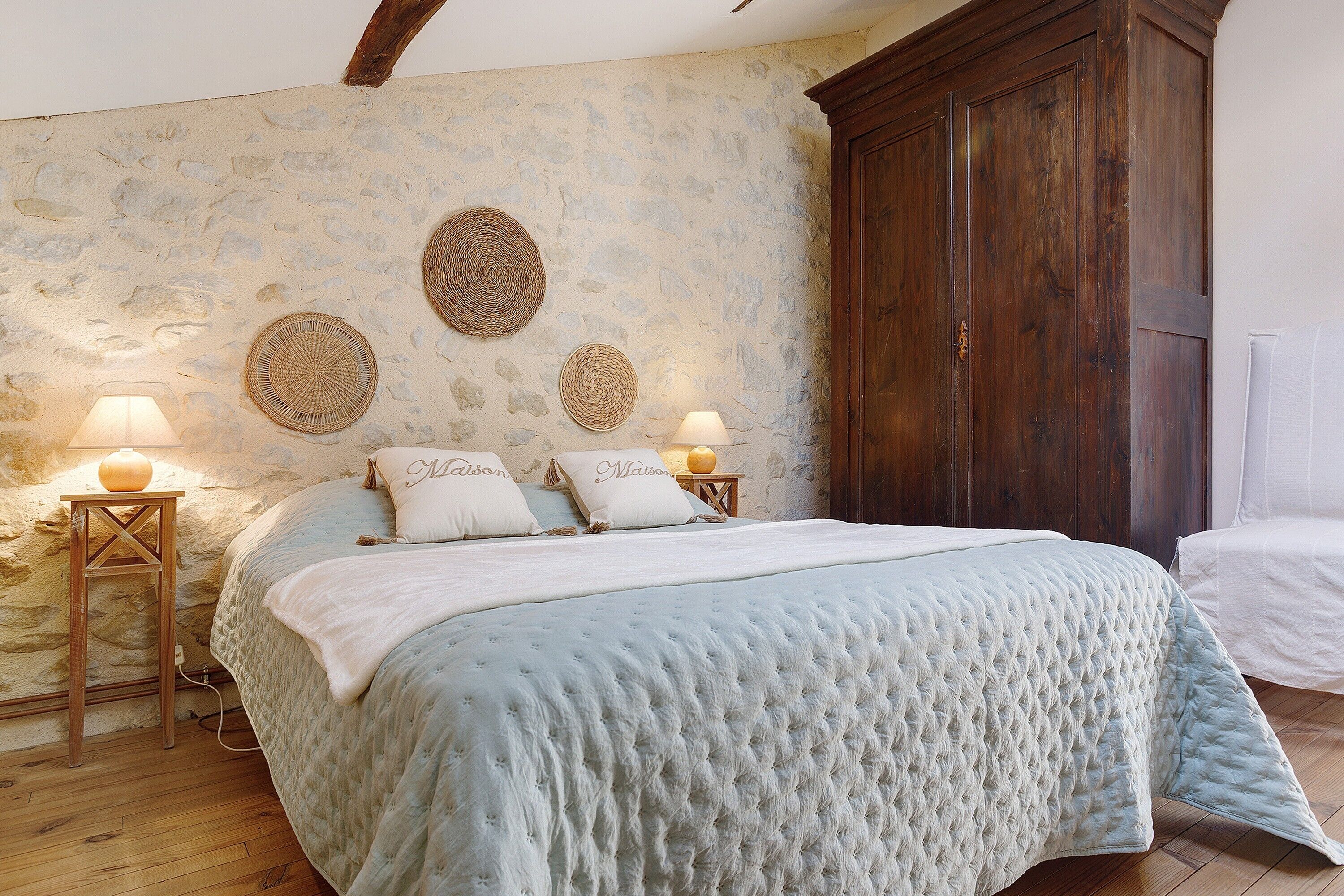 Un dormitorio con pared de piedra, un armario de madera, una cama con edredón acolchado y dos lámparas de noche.
