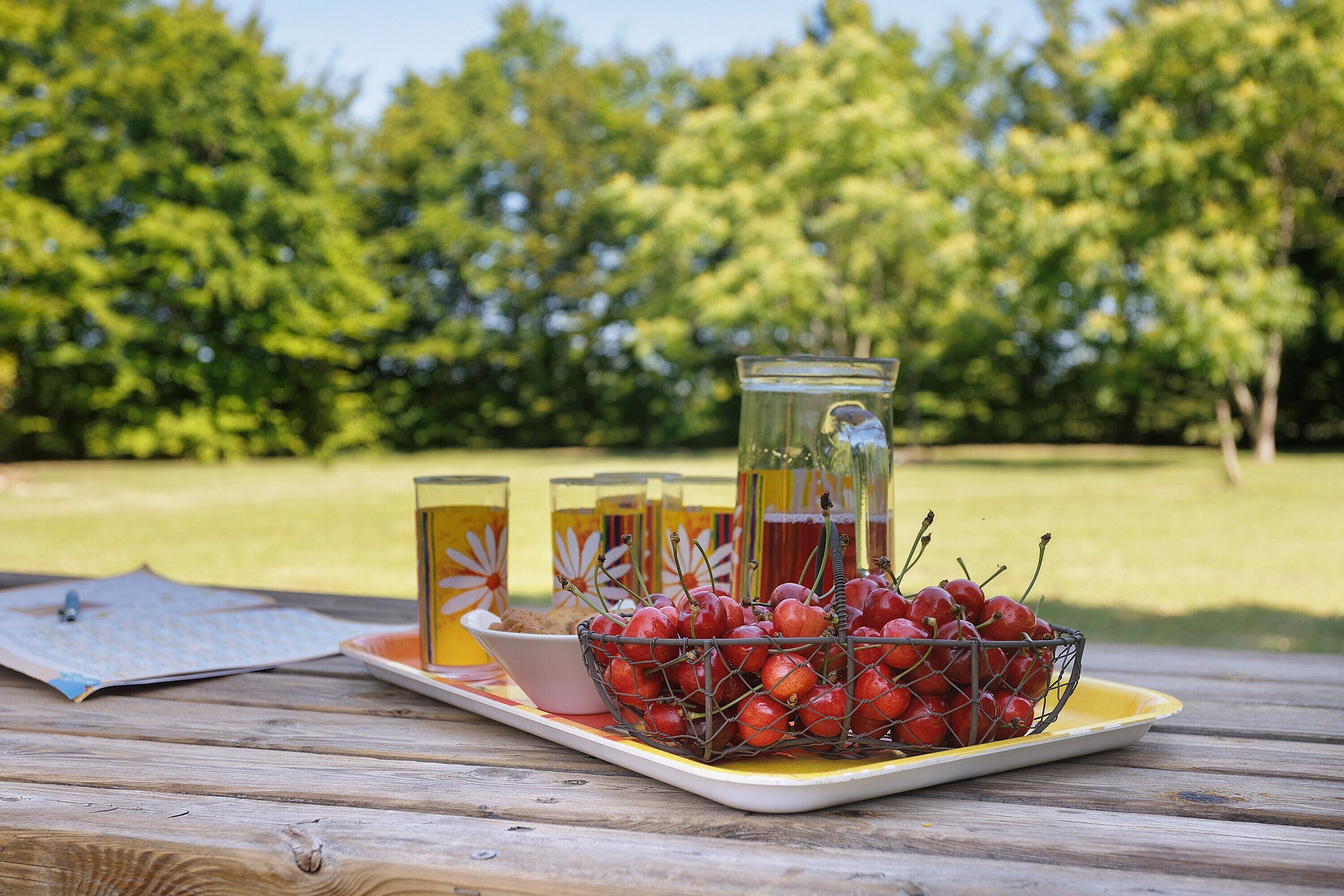 Une table en bois dressée pour un pique-nique, avec un panier de cerises, une carafe en verre et un plateau contenant des tasses.