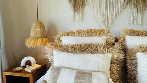 Quarto com cama, um pendente de macramé na parede, luminária trançada e mesa de cabeceira com luminária e objeto decorativo pequeno.