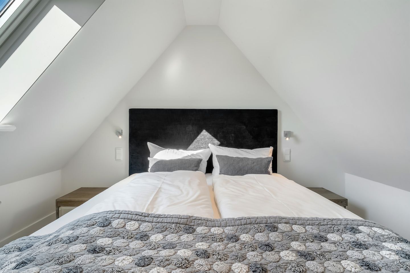 Quarto com teto inclinado, cama com roupa de cama branca, cabeceira preta e colcha estampada.