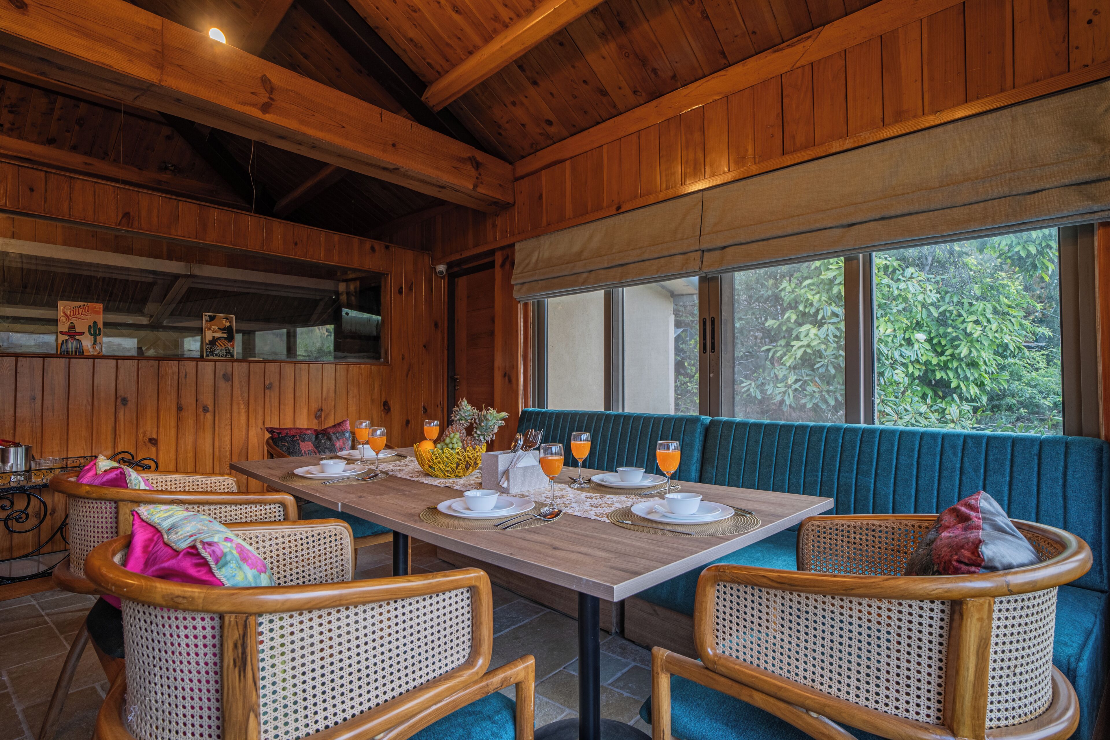 A wooden interior with a dining table set for four, surrounded by wicker chairs, and a window with a view of greenery.
