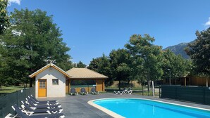 A swimming pool with lounge chairs, a small building, and a fenced area with trees and mountains in the background.