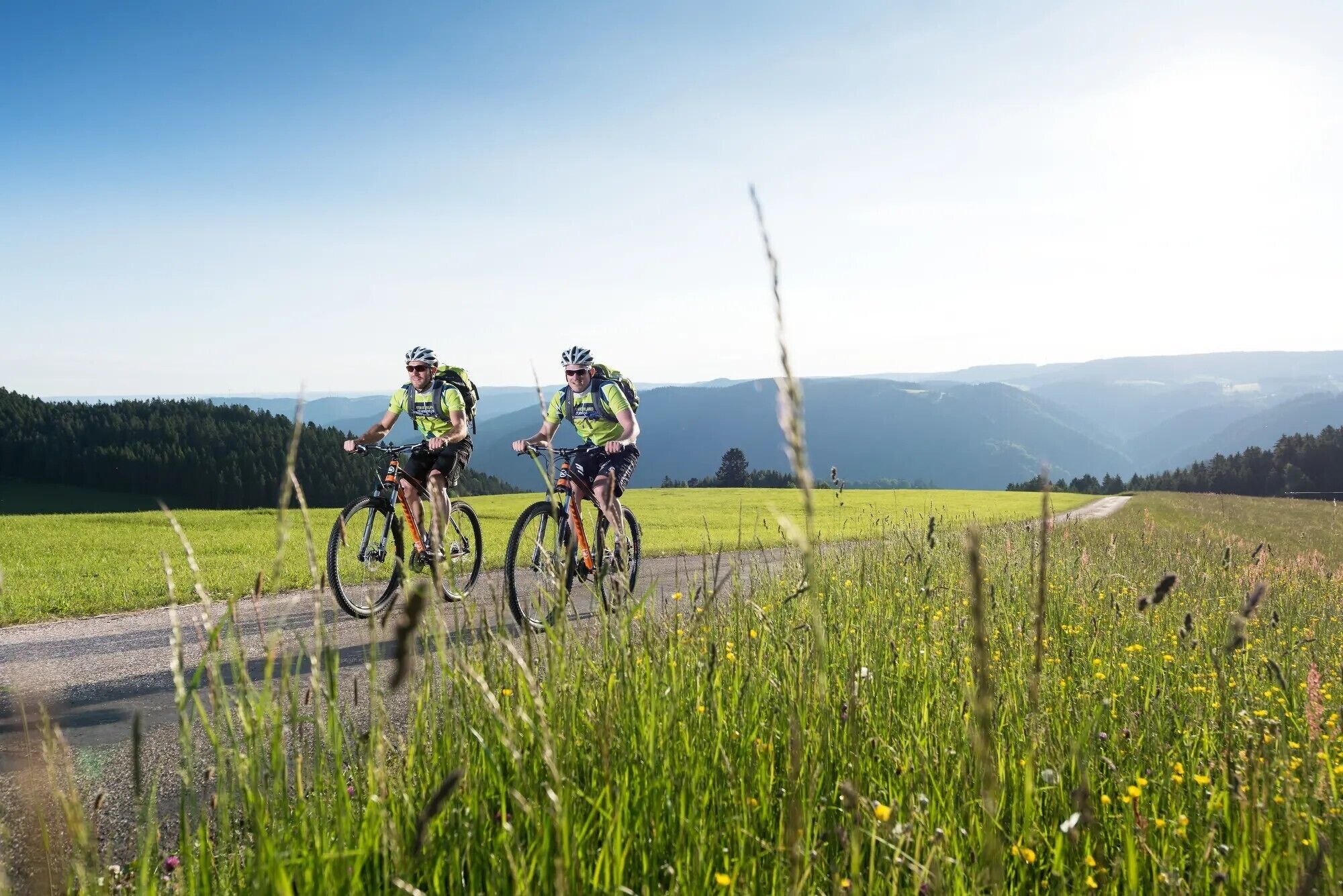 Two cyclists on a road with a scenic view of mountains and fields.