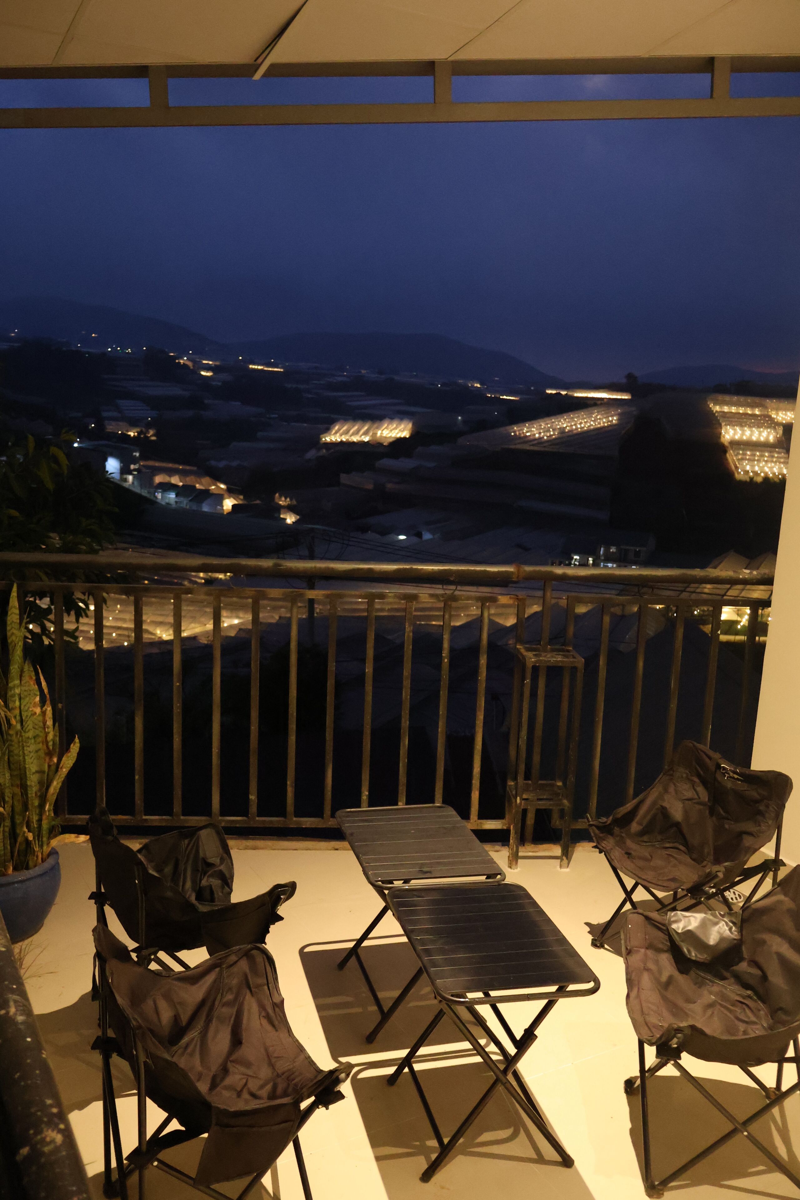 A balcony with a table and chairs, a potted plant, and a view of distant lights and mountains.