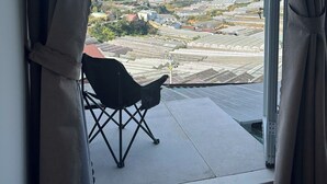Vista a través de una ventana con cortinas, un sillón y un balcón con cielo despejado y paisaje distante.