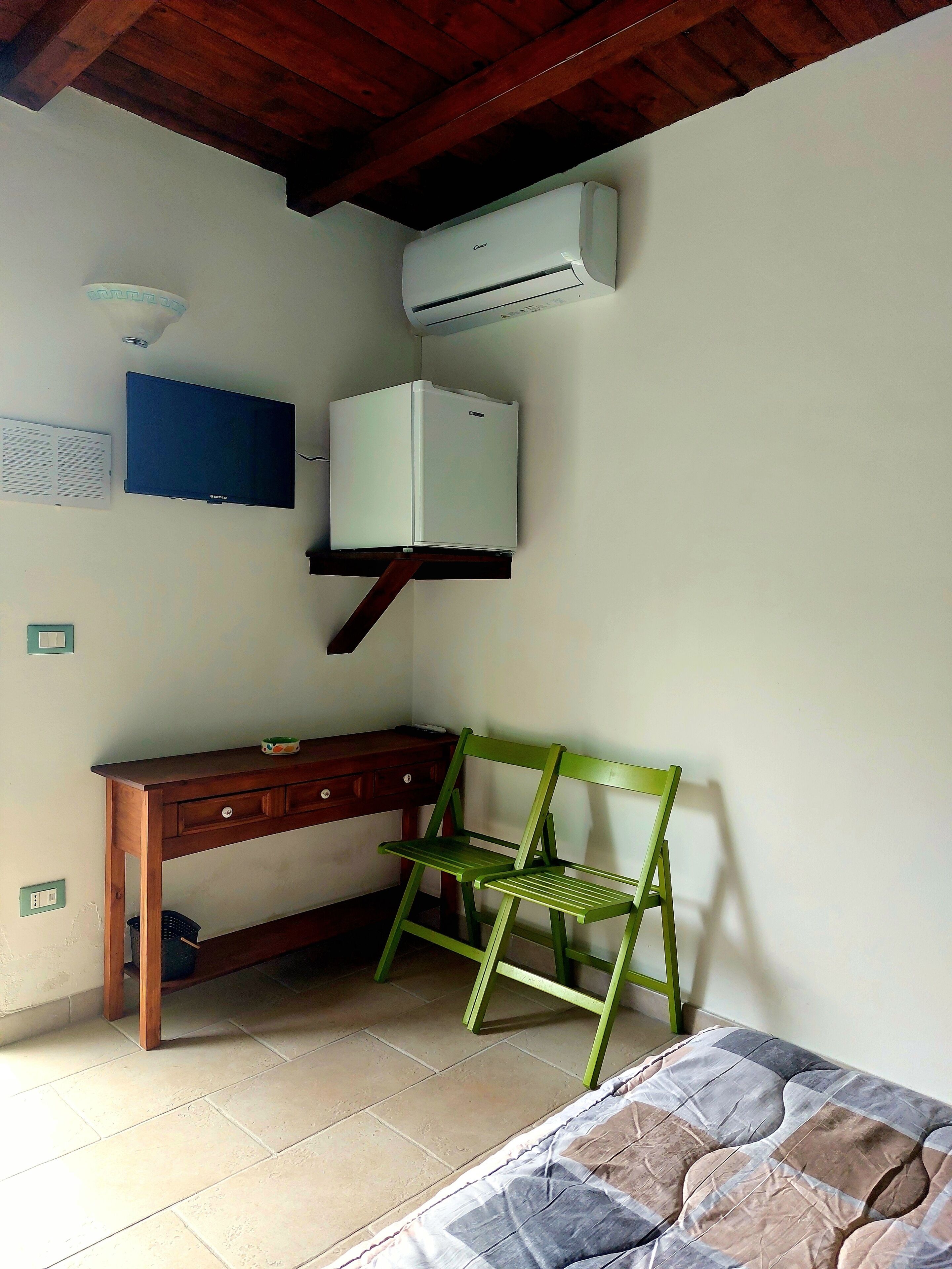 Une pièce comprenant un bureau en bois, une chaise verte, un lit avec une couvre-lit à motifs, un climatiseur et un téléviseur fixé au mur.