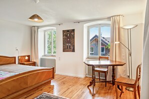 A bedroom with a wooden bed, a table, and a chair, a window with curtains, and a painting on the wall.