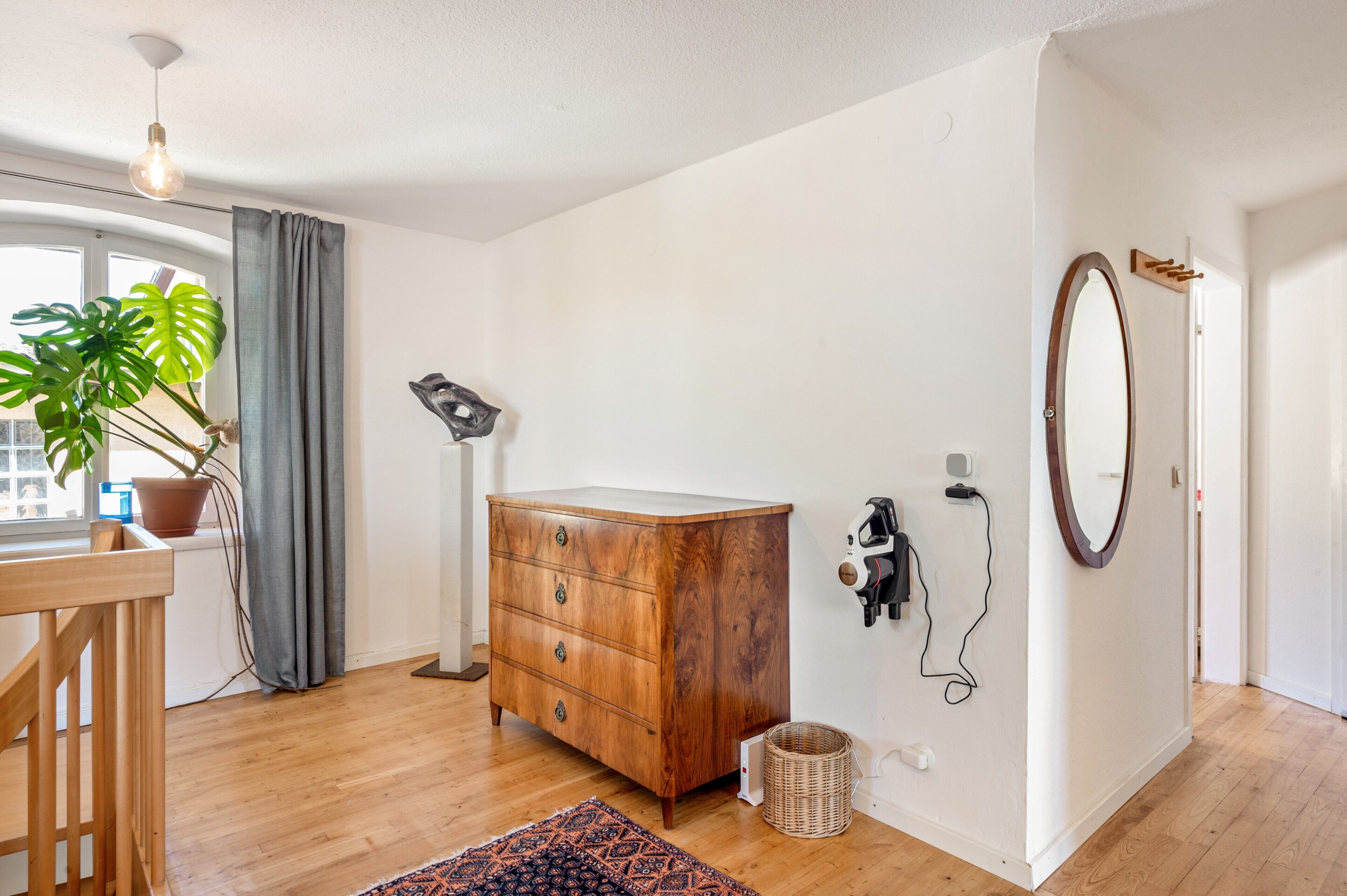 A room with a wooden dresser, a potted plant, a desk, a lamp, a mirror, and a wall-mounted speaker.