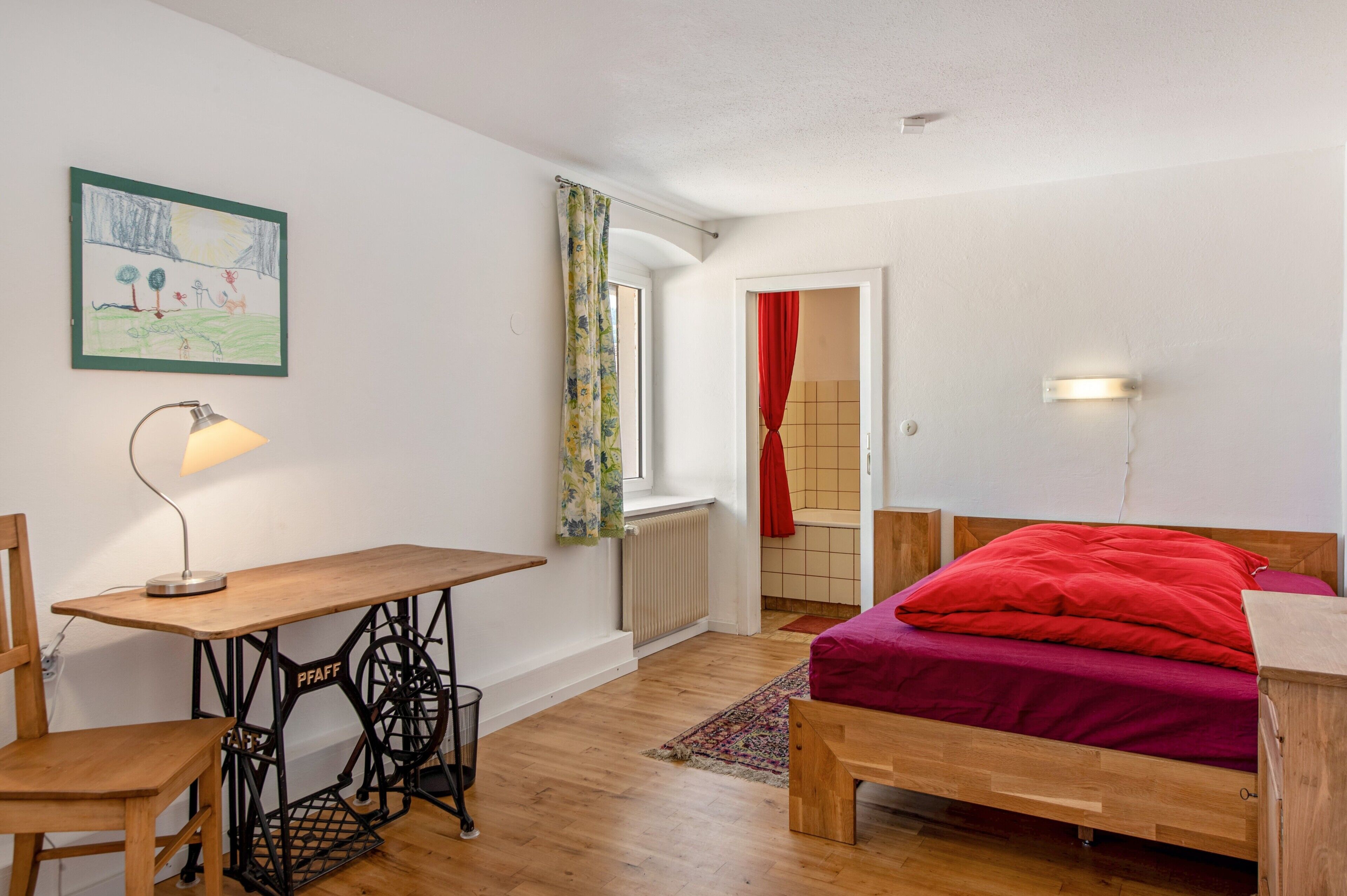 Ein Schlafzimmer mit einem hölzernen Bett, einem Schreibtisch mit Nähmaschine, einem Heizkörper und einem Fenster mit Vorhängen.