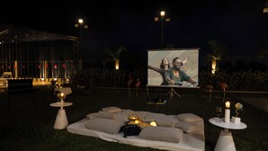 An outdoor seating area with a projector screen, tables with candles, and a cozy arrangement of cushions. - Alaya Stays Parinita Farm 4 BHK Villa (Faridabad)