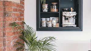 Ett rum med en tegelvägg, en krukväxt, en hylla med köksredskap och en kaffemaskin.