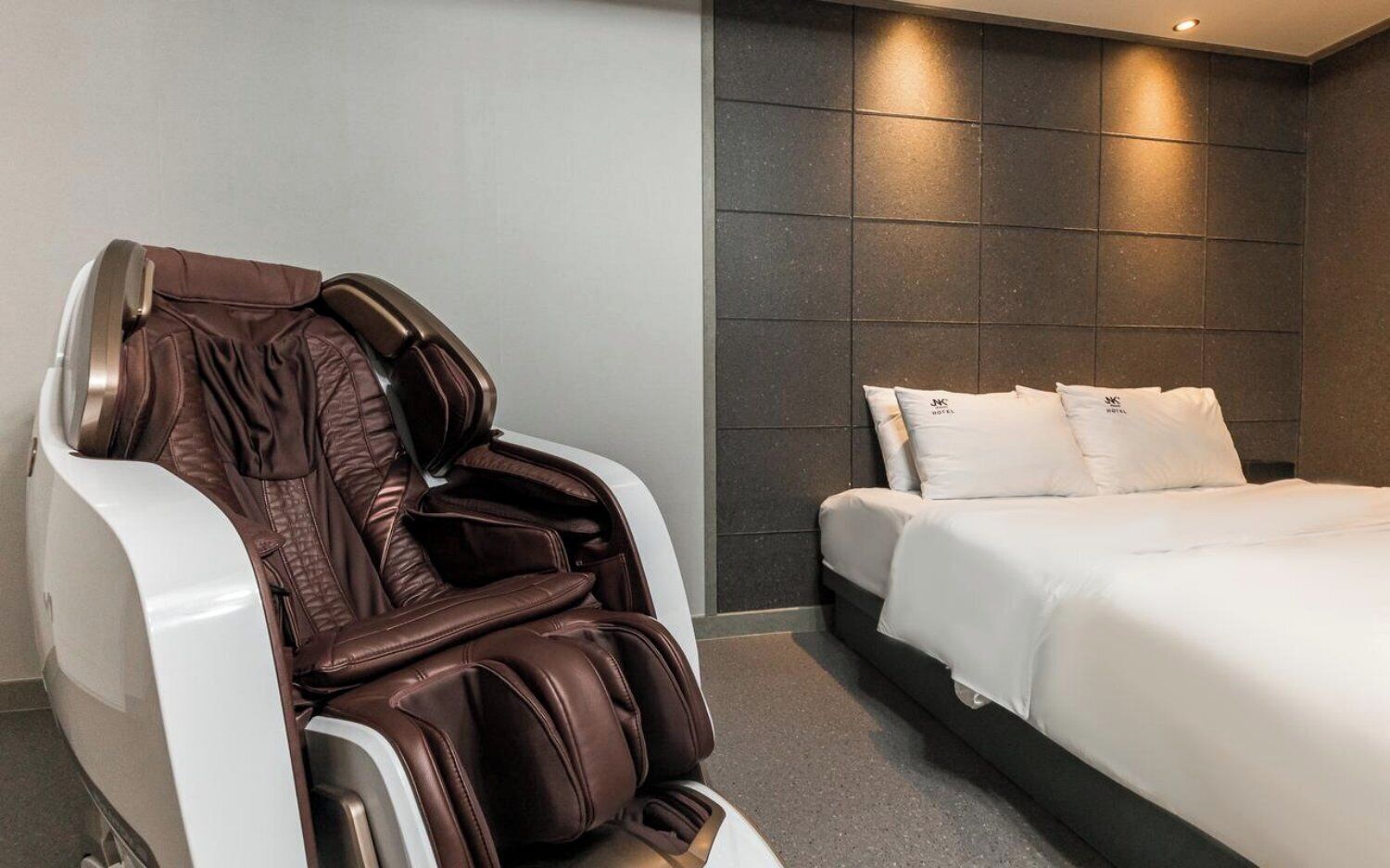 Una habitación de hotel moderna con una silla de masajes, una cama bien hecha y una pared de baldosas oscuras.