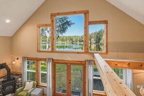 Una habitación con chimenea de piedra, un sillón verde y vistas a un lago a través de amplios ventanales.