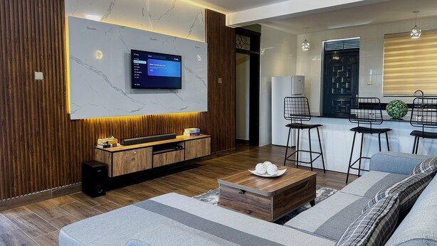A modern living room with a flat-screen TV, wooden paneling, a sofa, and a coffee table. - Vee Hotel Apartments (Entebbe)