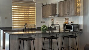 A modern kitchen with a central island, bar stools, wooden cabinets, and a window with blinds. - Vee Hotel Apartments (Entebbe)