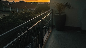 A balcony with a railing, a potted plant, and a view of the sunset over a residential area. - Vee Hotel Apartments (Entebbe)