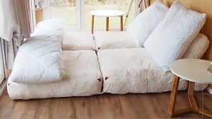 Un sofá modular con cojines blancos, una mesa redonda de madera y vistas al exterior a través de amplios ventanales.