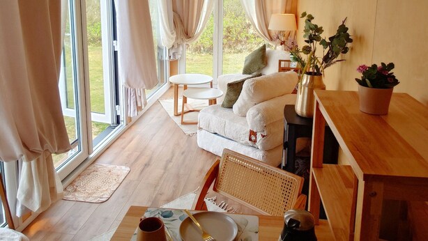 Una habitación acogedora con sofá, una mesa de comedor con posavasos, una cafetera y vistas al exterior a través de amplias ventanas.