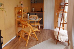 A compact room with a wooden dining table, two chairs, a small shelf with a lamp, and a ladder with folded clothes.