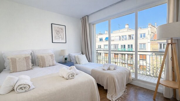 Een hotelkamer met twee bedden, een raam met uitzicht op gebouwen, een lijst met een afbeelding aan de muur, en een tafeltje met een lamp.