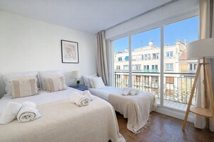Habitación de hotel con dos camas, una ventana con vista a edificios, un cuadro en la pared y una mesita con una lámpara.