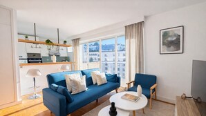 Een moderne woonkamer met een blauwe bank, twee witte ronde tafels, een stoel, een televisie en een keuken op de achtergrond.