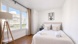 Een hotelkamer met een groot bed, een staande lamp, een nachtkastje met een lamp en uitzicht op gebouwen door het raam.