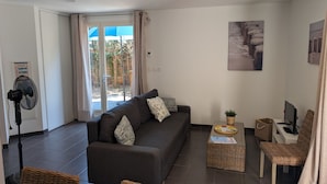 Ein Wohnzimmer mit einer Couch, zwei Korbstühlen, einem kleinen Tisch mit einer Pflanze, einem Ventilator und einem Fernseher.