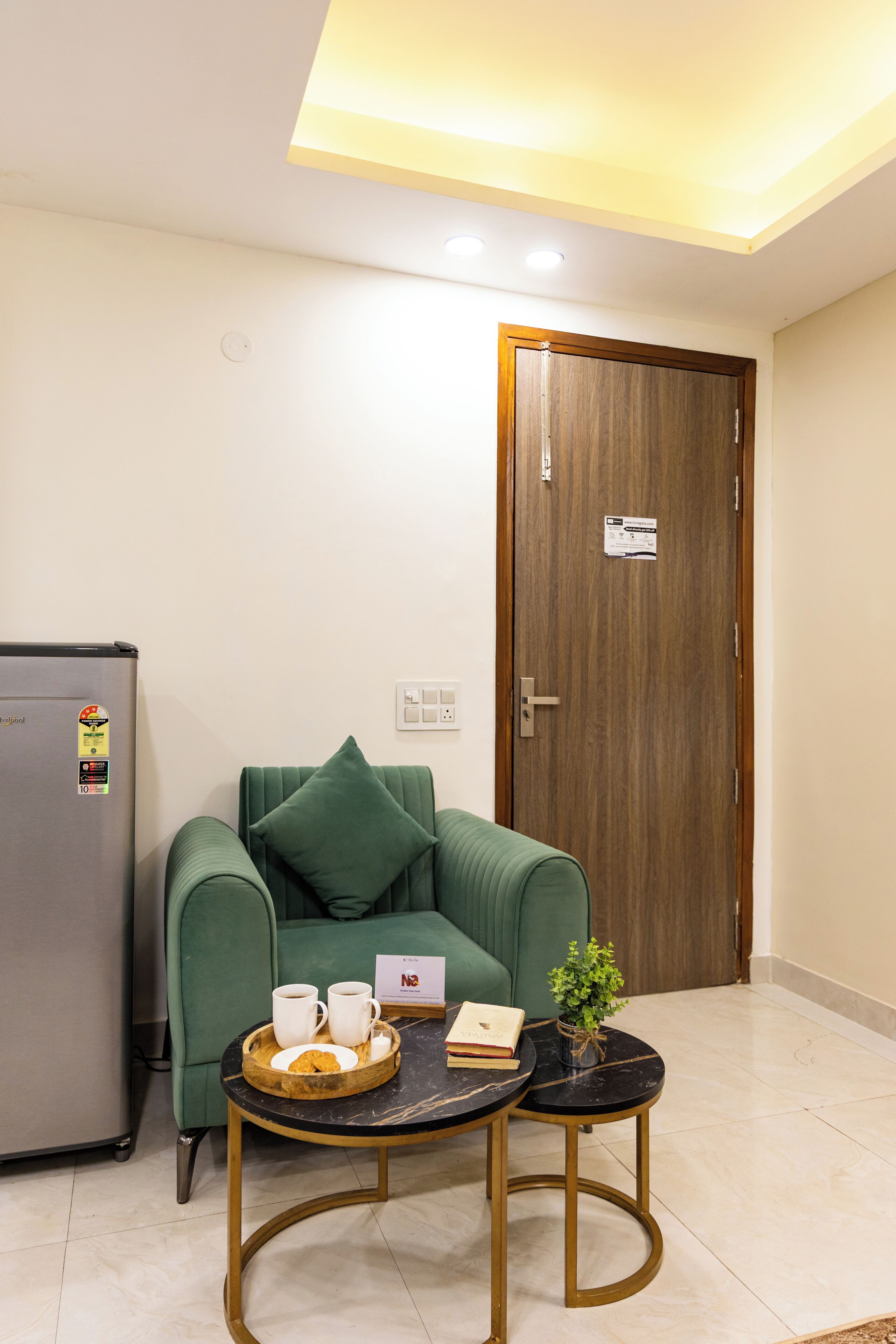 A room with a green sofa, a small round table with a tray of items, a refrigerator, and a wooden door.