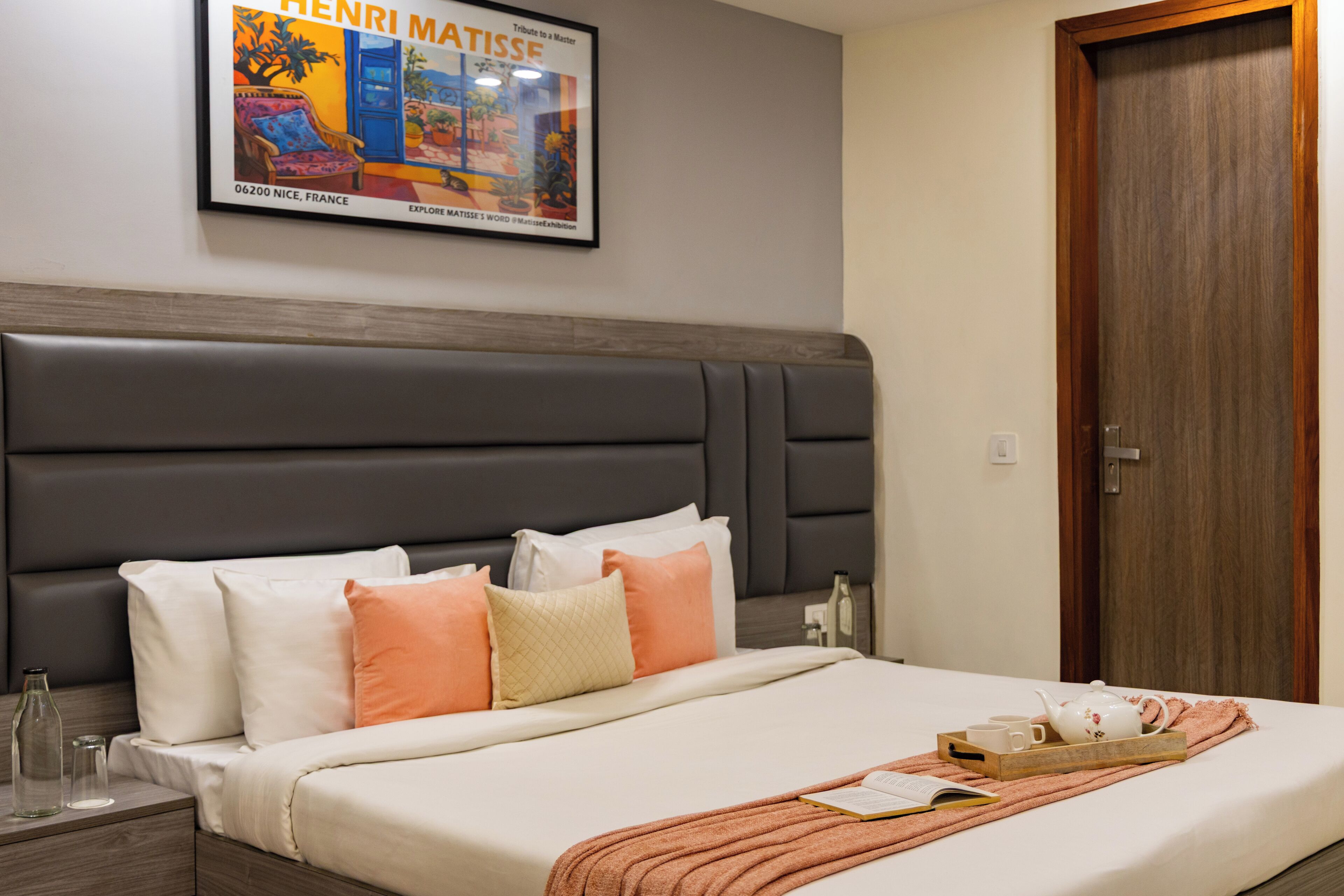 A neatly made bed with pillows, a bedside table with a bottle and glasses, a framed artwork on the wall, and a wooden door.
