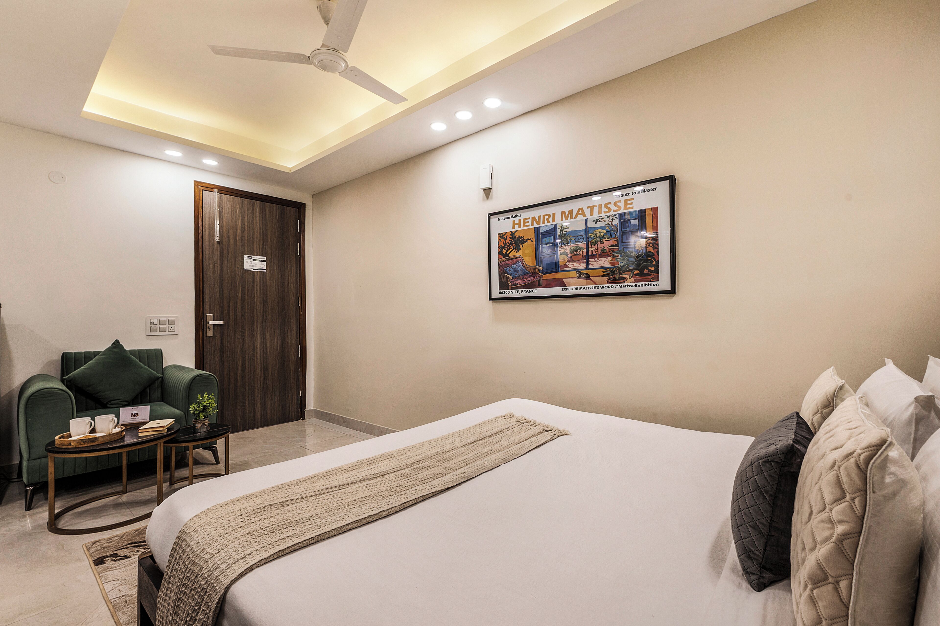 A hotel room with a bed, a desk, a chair, a ceiling fan, recessed lighting, and a framed poster on the wall.