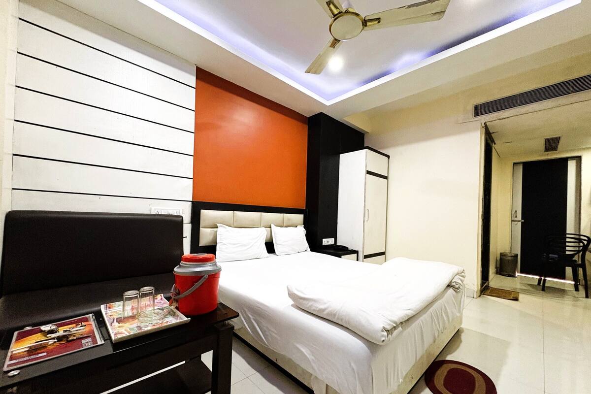 A hotel room with a bed, a nightstand, a red bucket, a ceiling fan, and a door leading to another room.