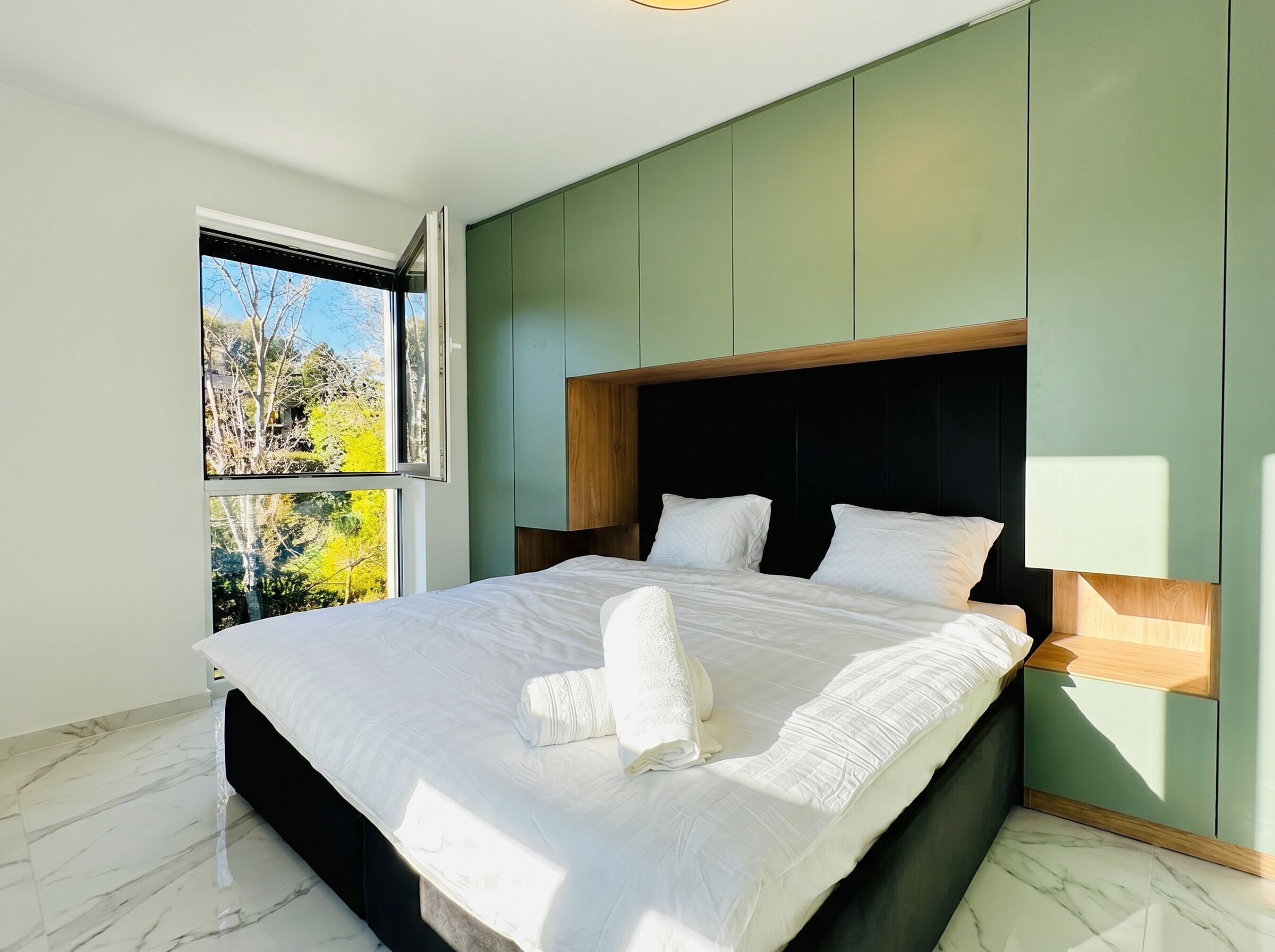 A modern bedroom with a large bed, green walls, a wooden headboard, and a window with a view of trees.