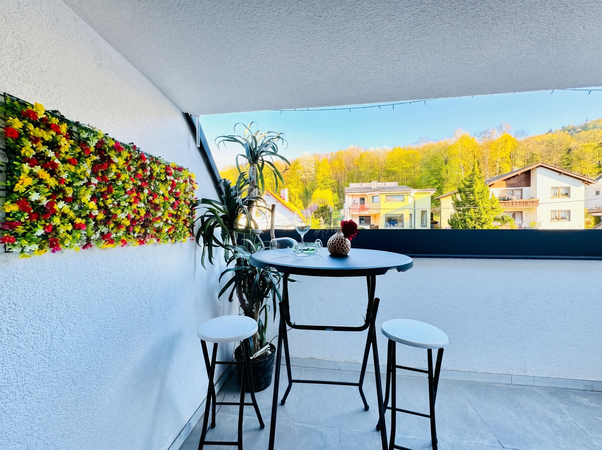 A balcony with a table, two chairs, a potted plant, and a wall of flowers.