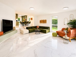 A modern living room with a flat-screen TV, green sofa, orange sofa, and a dining area with chairs and a table.