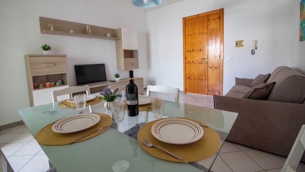 A dining area with a glass table, plates, cutlery, and a bottle of wine. There is a wooden door and a brown sofa in the background. - Charming Holiday House With Ocean Views in Otranto (Otranto)