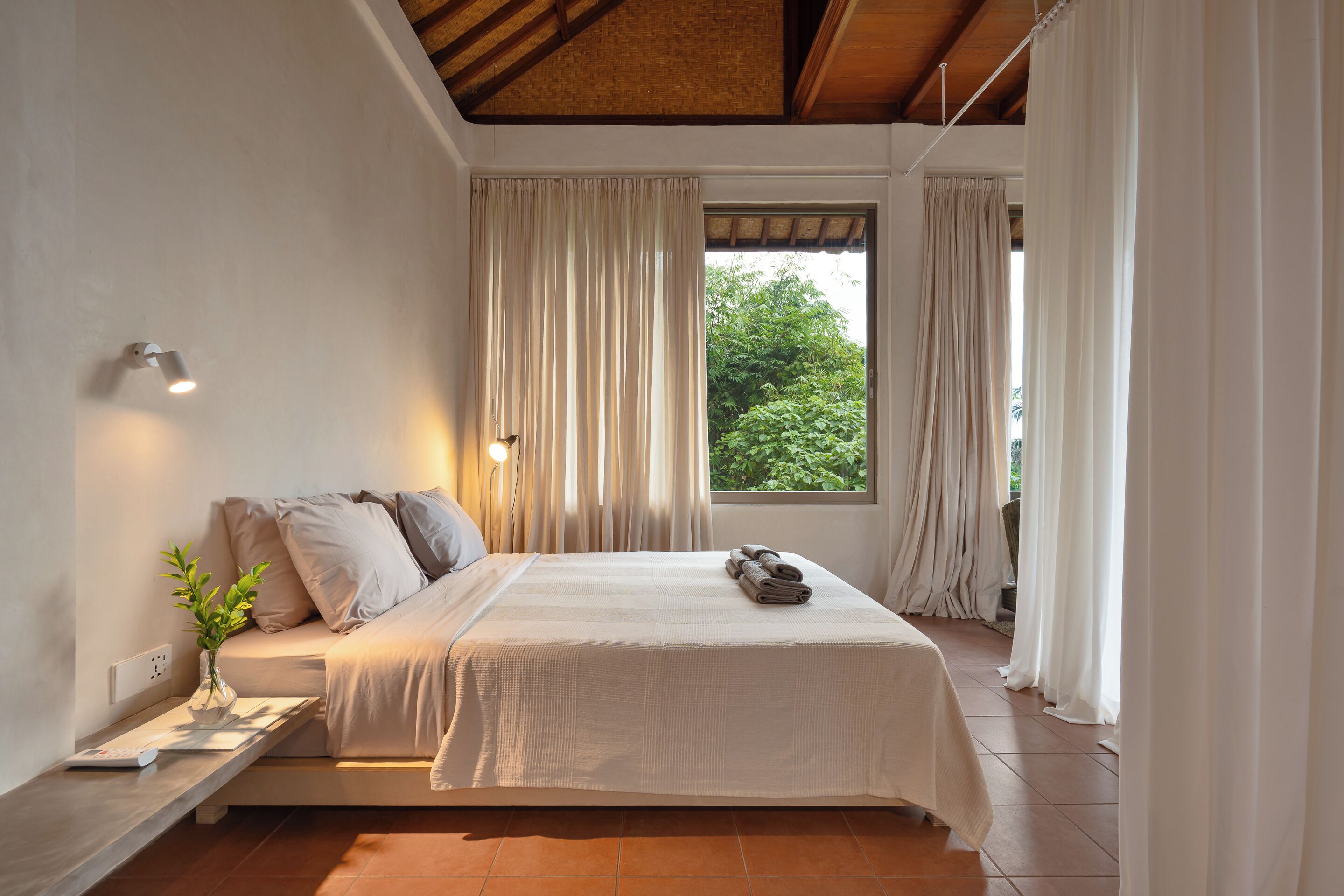 Une chambre à coucher avec un lit, une table de chevet avec une lampe, une fenêtre avec des rideaux et une vue sur la verdure à l’extérieur.