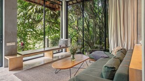 A modern living room with a sofa, bench, and coffee table, large windows with a view of greenery, and a concrete ceiling. - Pager Apartments 2BR in Ubud Center (Ubud)