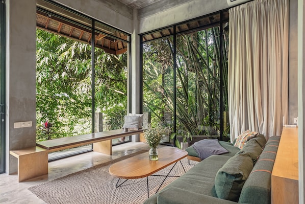 A modern living room with a sofa, bench, and coffee table, large windows with a view of greenery, and a concrete ceiling. - Pager Apartments 2BR in Ubud Center (Ubud)