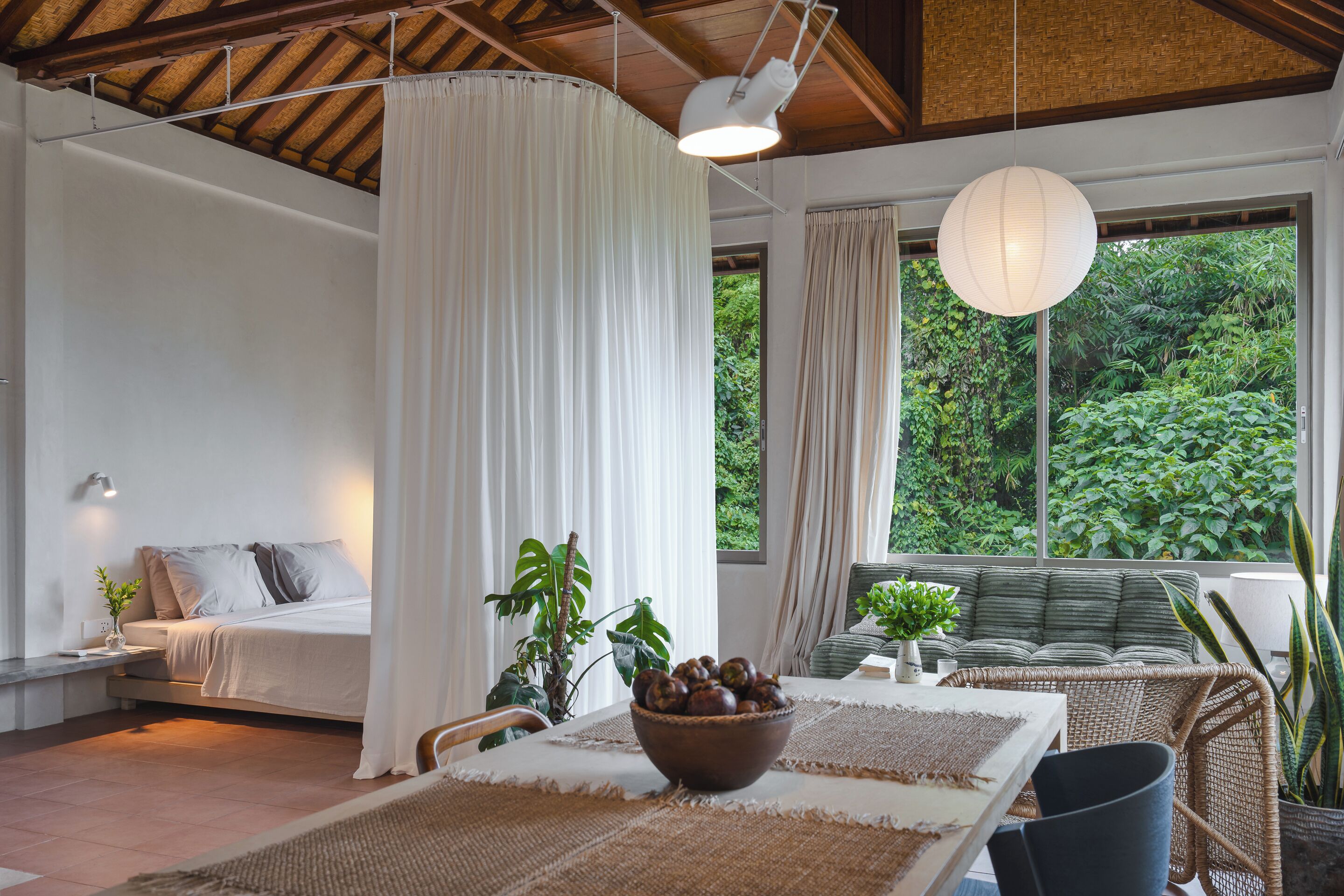 A room with a bed, a table with a bowl of fruit, and a view of greenery outside.