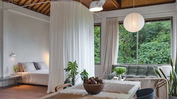 A room with a bed, a table with a bowl of fruit, and a view of greenery outside.