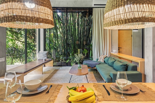A modern room with a sofa, dining table, and a view of greenery through large windows. - Pager Apartments - Nature View in City Center (Ubud)