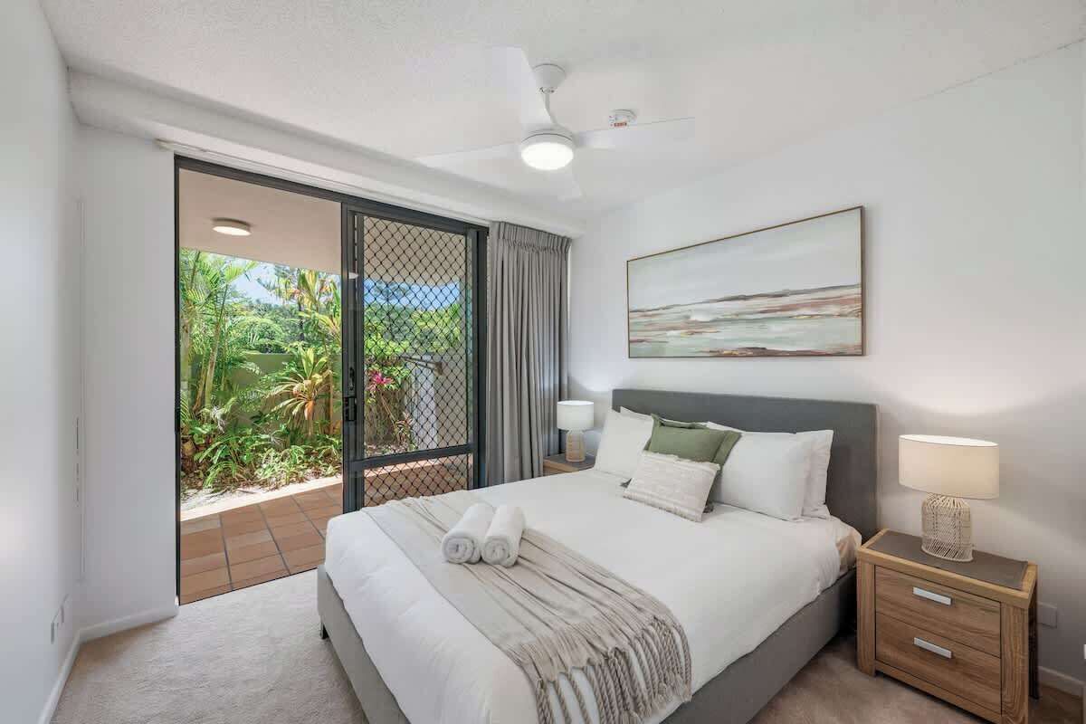 Une chambre à coucher comprenant un lit, une table de chevet, un ventilateur de plafond, une fenêtre donnant sur un paysage verdoyant et un tableau accroché au mur.